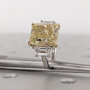 May include: A striking yellow diamond ring with a large, rectangular central stone. The ring has a gold setting around the main stone and smaller, rectangular diamonds on either side. The band is silver-coloured.