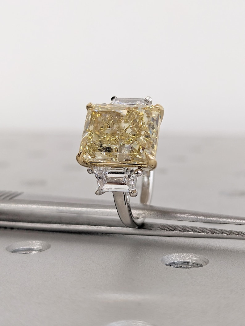 May include: A striking yellow diamond ring with a large, rectangular central stone. The ring has a gold setting around the main stone and smaller, rectangular diamonds on either side. The band is silver-coloured.
