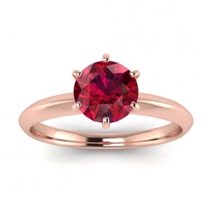 May include: A rose gold ring with a round, red ruby gemstone set in a six-prong setting.
