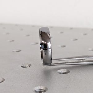 May include: A polished, silver-colored ring held by tweezers. The ring has a smooth, rounded surface and reflects light. The ring is a simple band design, suitable for various occasions.
