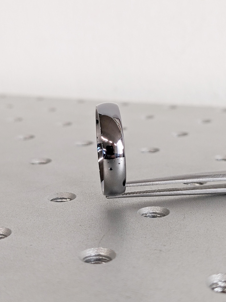May include: A polished, silver-colored ring held by tweezers. The ring has a smooth, rounded surface and reflects light. The ring is a simple band design, suitable for various occasions.