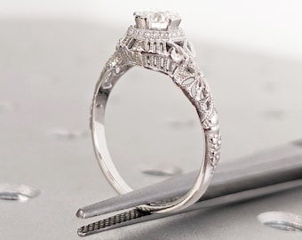 May include: A silver engagement ring with a round diamond set in a filigree design. The ring is held by a pair of tweezers.