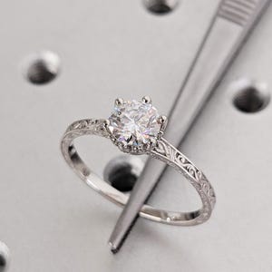 Pu&ograve; includere: Un anello di fidanzamento in argento con una grande gemma rotonda e trasparente. L'anello ha una fascia incisa ed &egrave; tenuto da una pinzetta. L'anello &egrave; posto su uno sfondo grigio chiaro.
