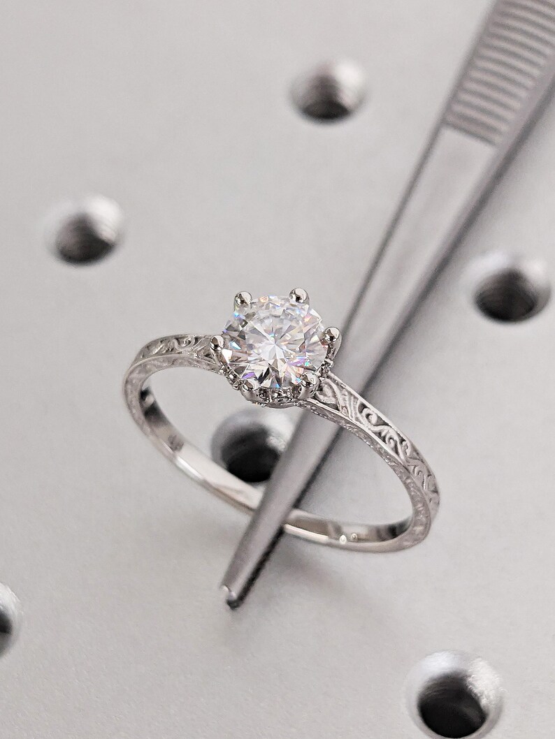 Pu&ograve; includere: Un anello di fidanzamento in argento con una grande gemma rotonda e trasparente. L'anello ha una fascia incisa ed &egrave; tenuto da una pinzetta. L'anello &egrave; posto su uno sfondo grigio chiaro.