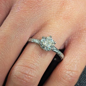 Pu&ograve; includere: Primo piano di un anello di fidanzamento in argento con una grande gemma rotonda e trasparente. L'anello ha una fascia ornata e incisa. L'anello &egrave; sul dito di una persona.