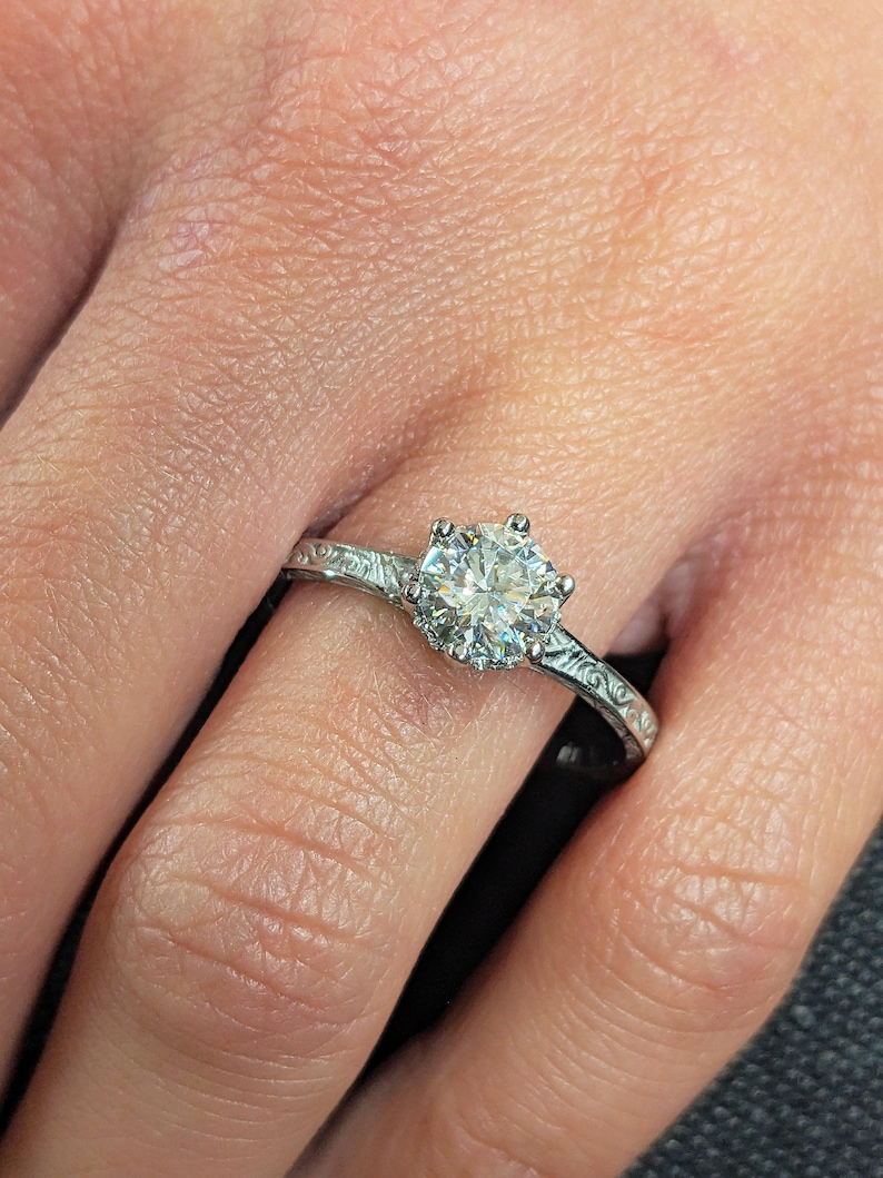 Pu&ograve; includere: Primo piano di un anello di fidanzamento in argento con una grande gemma rotonda e trasparente. L'anello ha una fascia ornata e incisa. L'anello &egrave; sul dito di una persona.