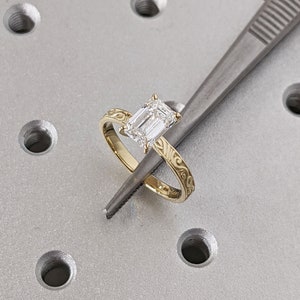 May include: A gold ring with an emerald cut diamond set in the center. The ring band is decorated with a delicate floral design.