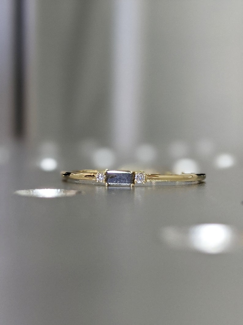 May include: A delicate gold band ring with a rectangular grey gemstone and two smaller clear gemstones on either side.
