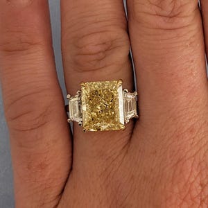 May include: A radiant-cut yellow diamond ring with two side diamonds. The large center stone is a vibrant yellow, set in a gold and platinum band. The ring's design features a classic three-stone setting, showcasing the brilliance of the diamonds.