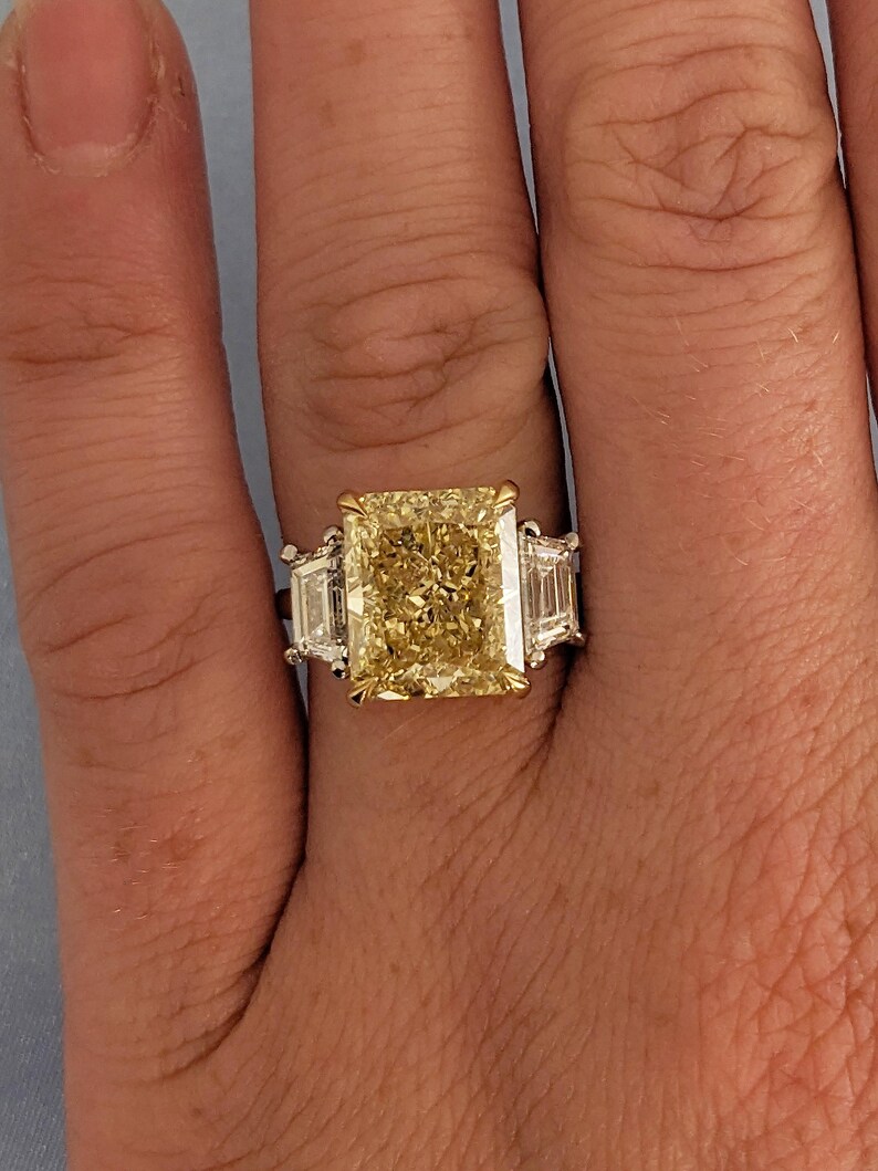 May include: A radiant-cut yellow diamond ring with two side diamonds. The large center stone is a vibrant yellow, set in a gold and platinum band. The ring's design features a classic three-stone setting, showcasing the brilliance of the diamonds.