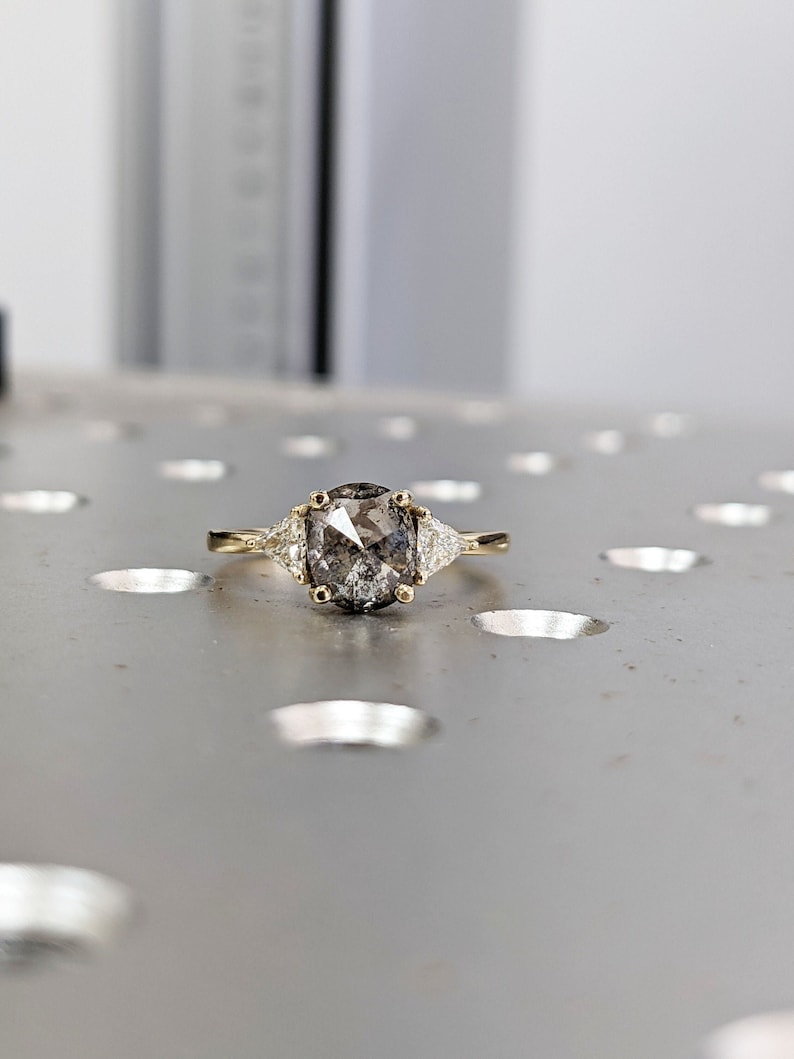 Puede incluir: Un anillo de compromiso de oro con un diamante gris ovalado y dos diamantes triangulares m&aacute;s peque&ntilde;os a cada lado.