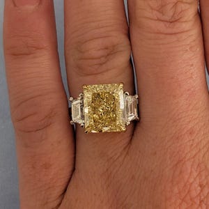 May include: A radiant-cut yellow diamond ring with two side stones. The large, central diamond is a vibrant yellow, set in a gold and silver band. The ring is a statement piece, perfect for special occasions.