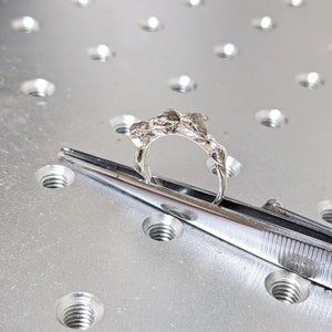 以下が含まれることがあります： テクスチャ加工が施された有機的なデザインのシルバーリング。リングは金属製の表面にピンセットで挟まれています。