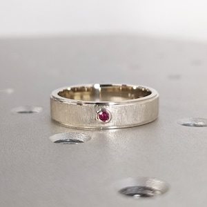 Puede incluir: Un anillo de banda de plata con una pequeña piedra roja engastada en el centro.