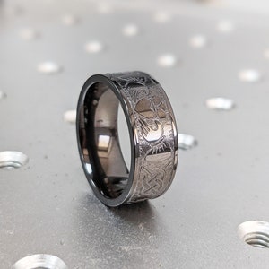 May include: A black metal ring featuring a Celtic knot design. The ring is engraved with a pattern of hearts and other symbols.