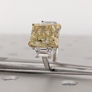 May include: A radiant-cut yellow diamond ring with a gold and silver band. The large, rectangular center stone is flanked by two smaller, rectangular diamonds. The ring is set on a silver band with gold accents, showcasing a luxurious design.