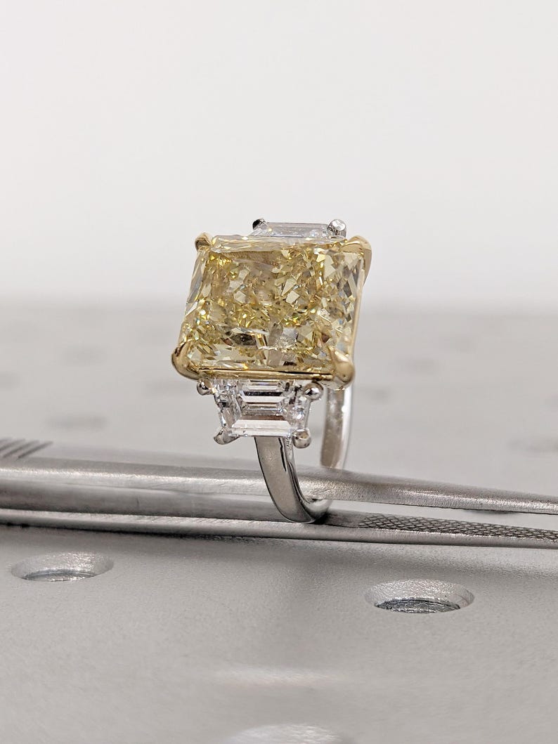 May include: A radiant-cut yellow diamond ring with a gold and silver band. The large, rectangular center stone is flanked by two smaller, rectangular diamonds. The ring is set on a silver band with gold accents, showcasing a luxurious design.