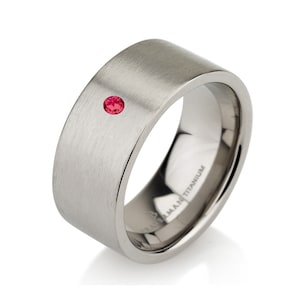 May include: A brushed titanium ring with a single, small, round, red gemstone. The ring has a flat, wide band and a minimalist design. The ring is a piece of jewelry.