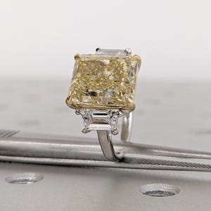 May include: A radiant, yellow diamond ring with a large, rectangular center stone. The ring features a gold setting around the main stone and smaller, rectangular diamonds on either side. The band is silver-colored.