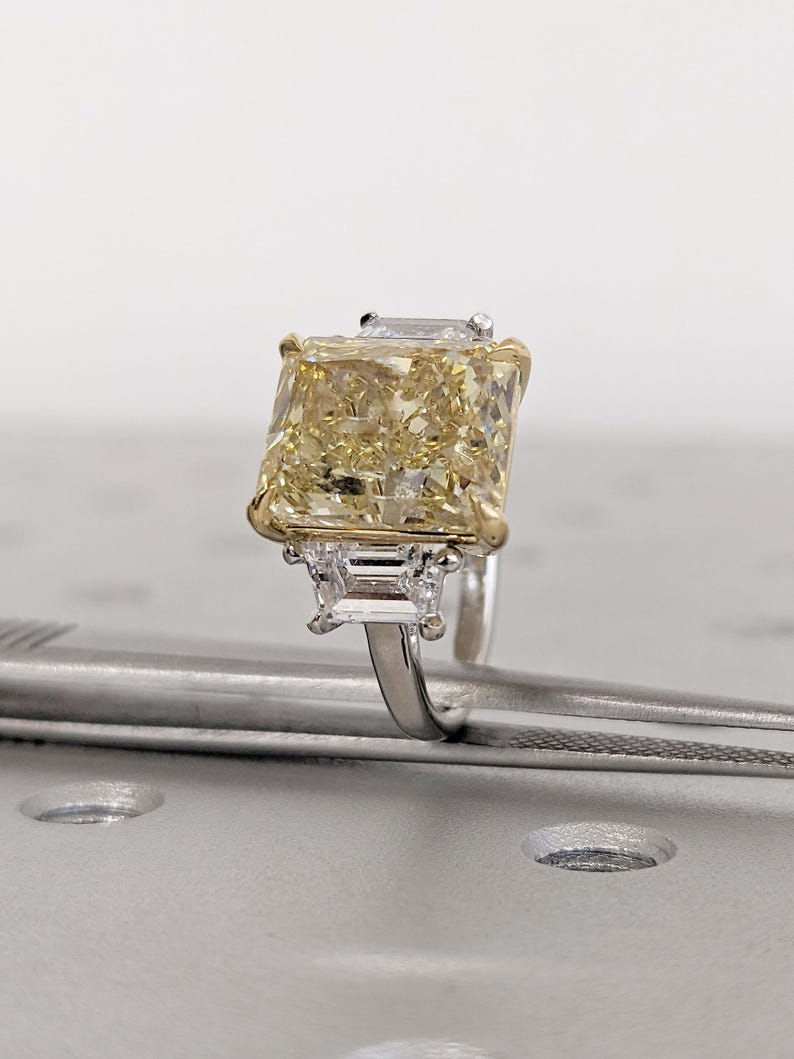 May include: A radiant, yellow diamond ring with a large, rectangular center stone. The ring features a gold setting around the main stone and smaller, rectangular diamonds on either side. The band is silver-colored.