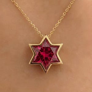 May include: A gold-colored necklace featuring a pendant in the shape of a six-pointed star. The star is filled with a faceted, deep red gemstone. The chain is delicate and also gold-colored, creating a striking contrast with the red gem.