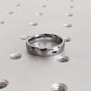 May include: A silver-colored ring with a smooth, polished surface. The ring is circular and has a simple, classic design. The ring is likely made of metal and is displayed on a light gray surface with evenly spaced holes.