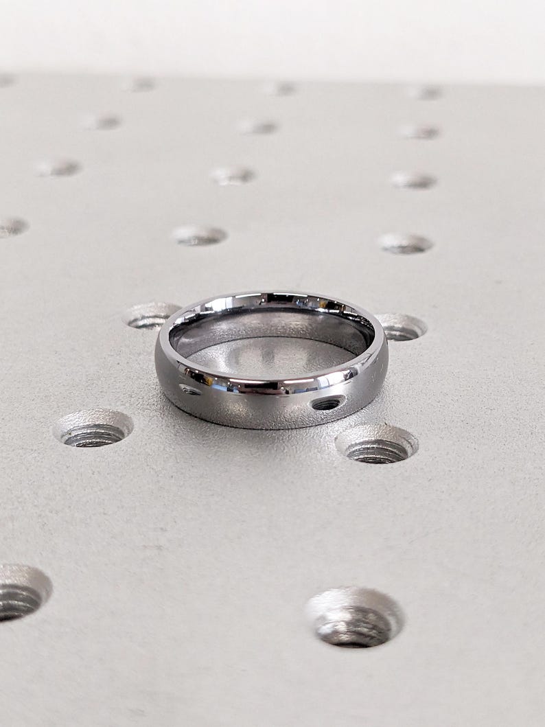 May include: A silver-colored ring with a smooth, polished surface. The ring is circular and has a simple, classic design. The ring is likely made of metal and is displayed on a light gray surface with evenly spaced holes.