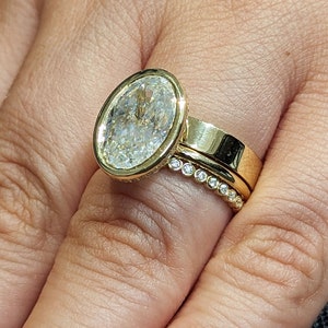 K&ouml;nnte beinhalten: Ein goldener Ring mit einem gro&szlig;en ovalen Diamanten, der in einer Zargenfassung sitzt. Der Ring wird an einem Finger mit einem zweiten goldenen Band getragen, das mit kleinen Diamanten in Pav&eacute;-Fassung besetzt ist.