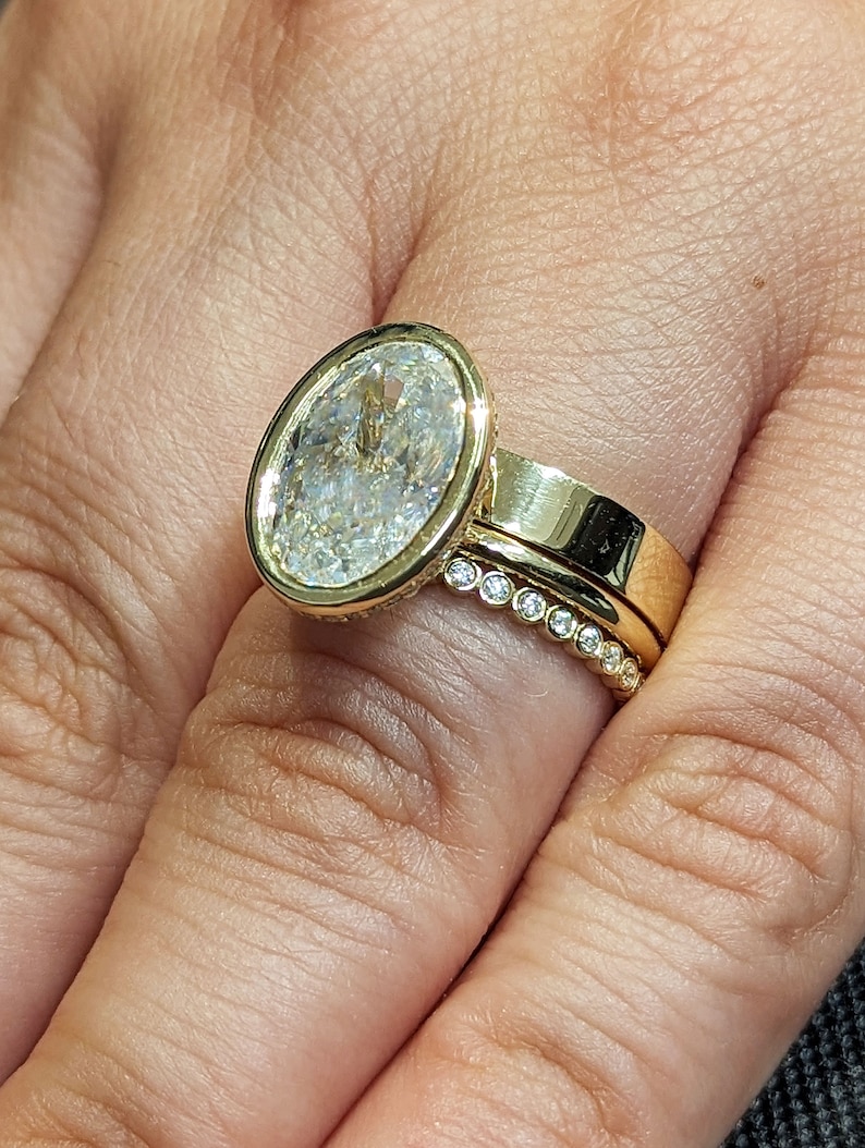 K&ouml;nnte beinhalten: Ein goldener Ring mit einem gro&szlig;en ovalen Diamanten, der in einer Zargenfassung sitzt. Der Ring wird an einem Finger mit einem zweiten goldenen Band getragen, das mit kleinen Diamanten in Pav&eacute;-Fassung besetzt ist.