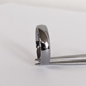 May include: A polished, dark gray ring with a smooth, rounded surface. The ring is held by a pair of silver tweezers, showcasing its reflective quality. The ring's design is simple and elegant, suitable for everyday wear.