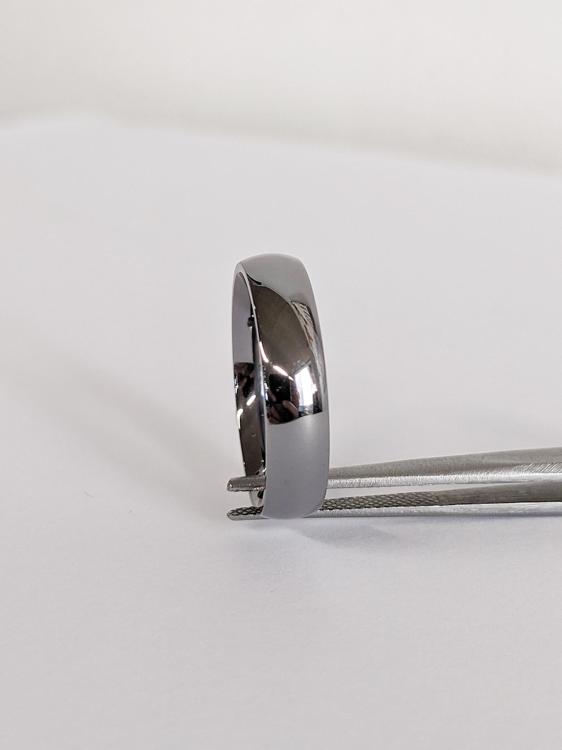 May include: A polished, dark gray ring with a smooth, rounded surface. The ring is held by a pair of silver tweezers, showcasing its reflective quality. The ring's design is simple and elegant, suitable for everyday wear.