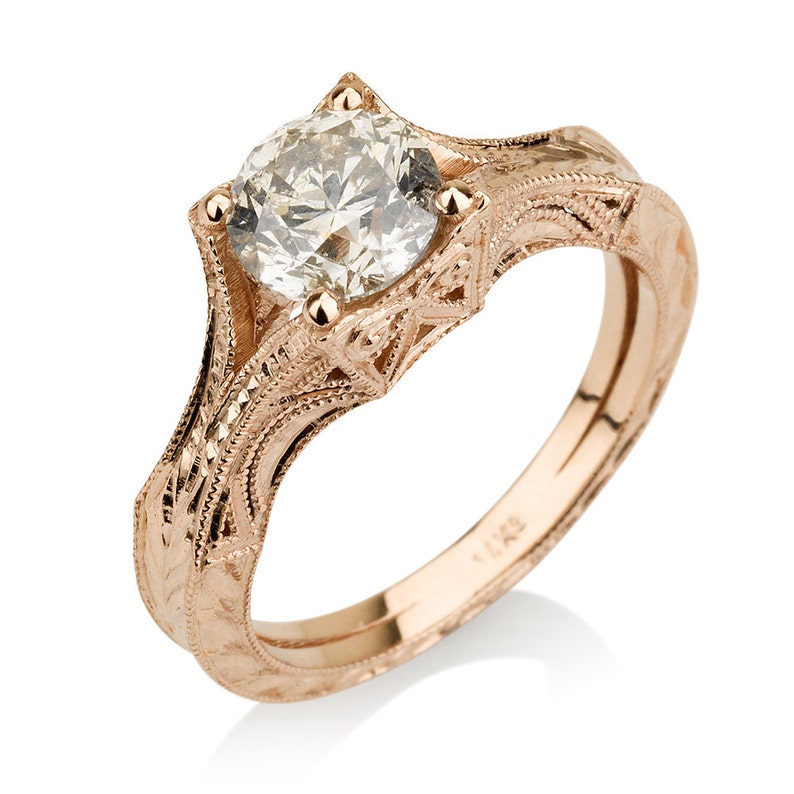 K&ouml;nnte beinhalten: Ein Verlobungsring aus Ros&eacute;gold mit einem runden Diamanten, der in einem filigranen Design gefasst ist. Die Band ist mit der Nummer "14K" graviert.