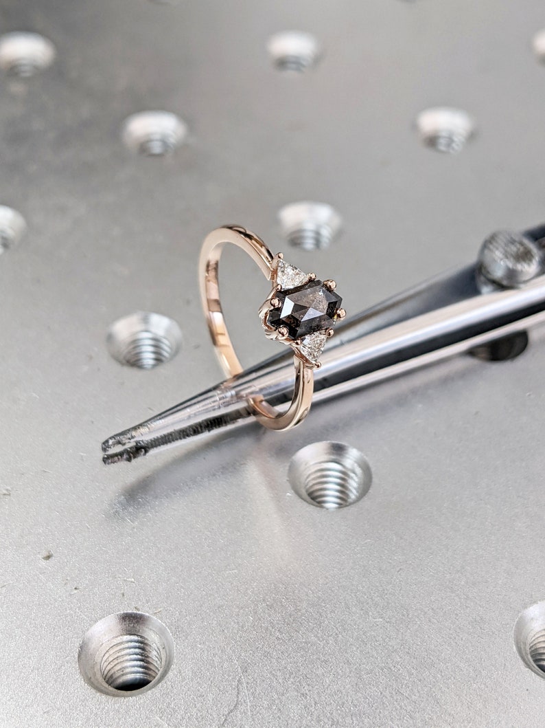 May include: A rose gold engagement ring with a black diamond center stone and three smaller white diamonds on the band.