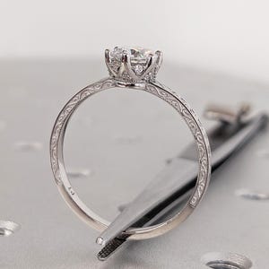 Pu&ograve; includere: Primo piano di un anello di fidanzamento in argento con una grande gemma trasparente. L'anello presenta una fascia ornata e incisa e un'incastonatura dettagliata. L'anello &egrave; tenuto da una pinzetta.