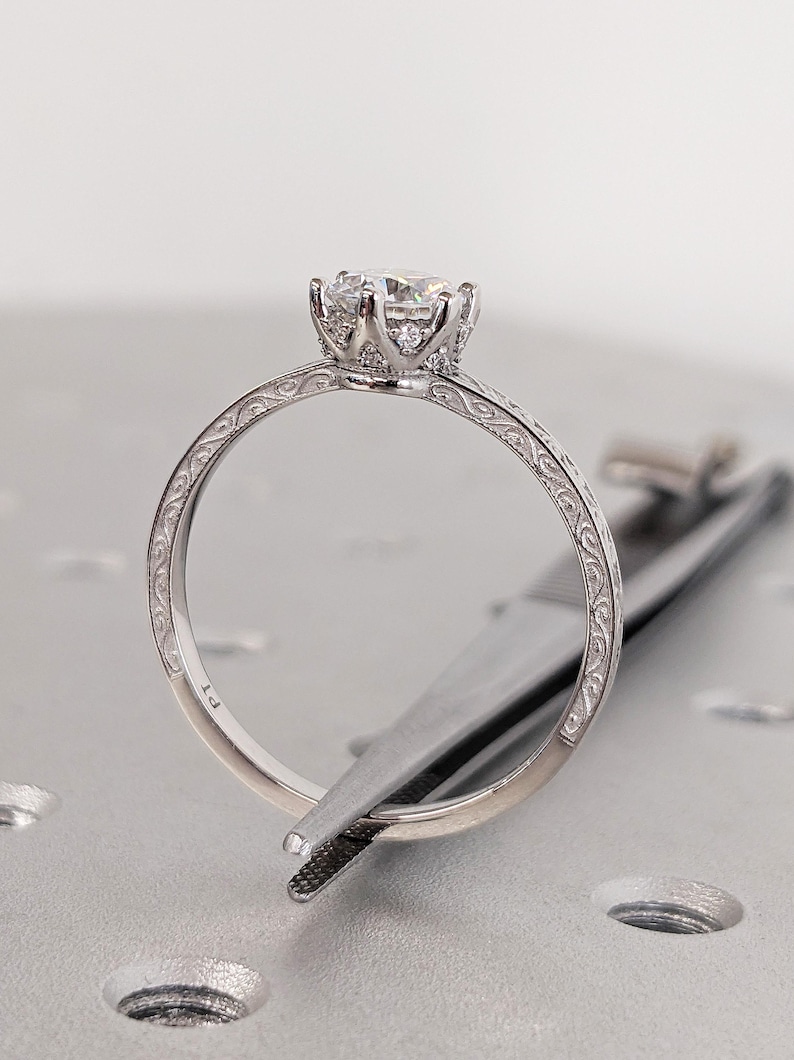 Pu&ograve; includere: Primo piano di un anello di fidanzamento in argento con una grande gemma trasparente. L'anello presenta una fascia ornata e incisa e un'incastonatura dettagliata. L'anello &egrave; tenuto da una pinzetta.