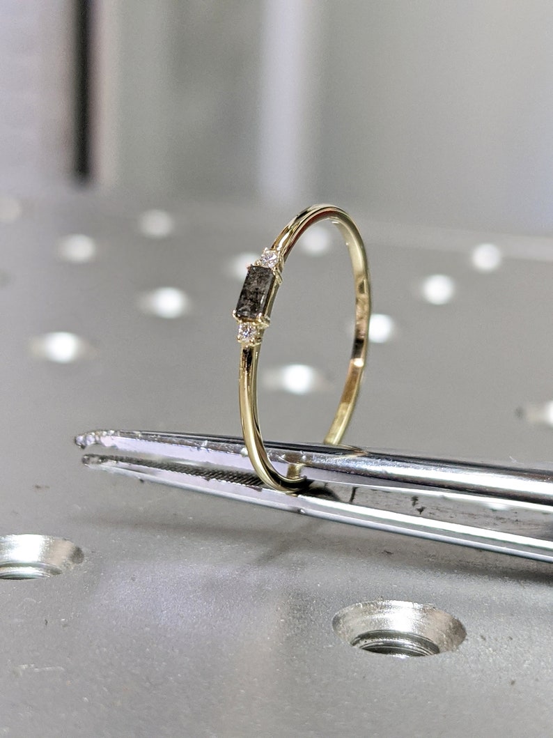 May include: A delicate gold band ring with a small, rectangular, grey diamond and two smaller clear diamonds.