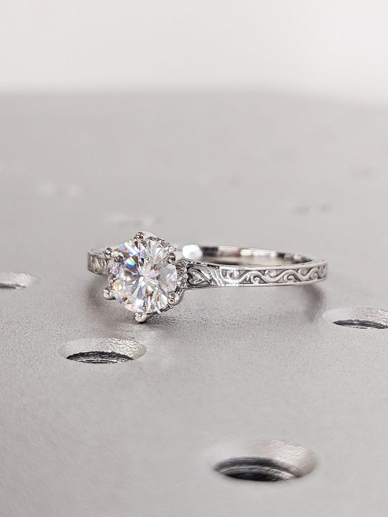 Pu&ograve; includere: Un anello di fidanzamento in argento con una grande gemma rotonda e trasparente. L'anello ha una fascia ornata e incisa e un'incastonatura a sei punte. L'anello &egrave; esposto su una superficie grigio chiaro.