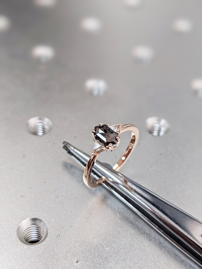 May include: A rose gold engagement ring with a black diamond center stone and two smaller diamonds on the side. The ring is held by tweezers on a gray surface.