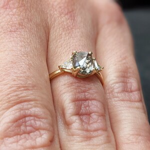 Puede incluir: Un anillo de oro con un gran diamante gris de forma ovalada rodeado de dos diamantes triangulares m&aacute;s peque&ntilde;os.