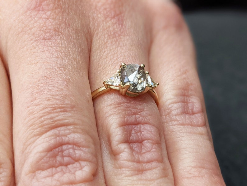 Puede incluir: Un anillo de oro con un gran diamante gris de forma ovalada rodeado de dos diamantes triangulares m&aacute;s peque&ntilde;os.