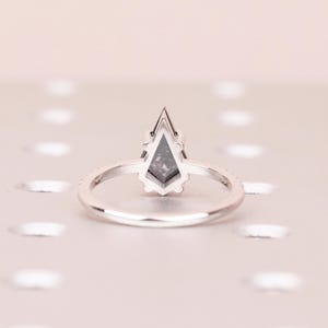 Puede incluir: Un anillo de plata con una piedra preciosa gris en forma de cometa. La banda es delgada y presenta peque&ntilde;as piedras transparentes. El anillo est&aacute; sobre un fondo rosa claro, creando una est&eacute;tica minimalista.