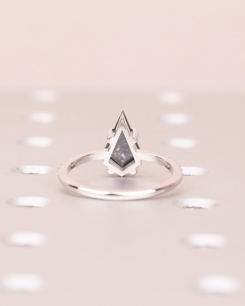 Puede incluir: Un anillo de plata con una piedra preciosa gris en forma de cometa. La banda es delgada y presenta peque&ntilde;as piedras transparentes. El anillo est&aacute; sobre un fondo rosa claro, creando una est&eacute;tica minimalista.