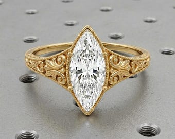 May include: A gold marquise-cut engagement ring with a large, clear gemstone. The ring features intricate filigree detailing along the band and around the gemstone setting. The ring is set against a neutral background.