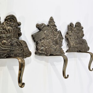 May include: Three vintage metal stocking holders featuring Santa Claus in a sleigh, Santa Claus with a Christmas tree, and Santa Claus with a house. The holders are all in a bronze color and have a hook on the bottom.