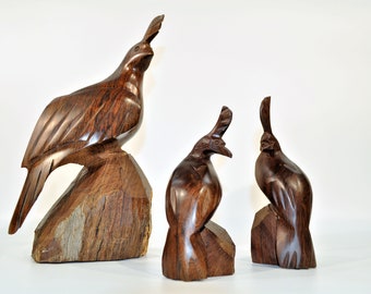 May include: Three wooden carvings of quail birds. The largest carving is of a quail standing on a rock, with its head tilted up. The two smaller carvings are of quail sitting on rocks, with their heads tilted down. The carvings are all made of dark brown wood.