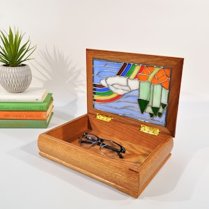 May include: A wooden box with a stained glass lid depicting a rainbow, clouds, and pencils. The box is open and a pair of glasses are inside.