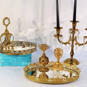 Puede incluir: Un conjunto de tres bandejas decorativas doradas con detalles ornamentados. Las bandejas presentan superficies espejadas y son perfectas para exhibir objetos peque&ntilde;os o joyas. Las bandejas est&aacute;n dispuestas sobre una superficie blanca con un candelero dorado en el fondo.
