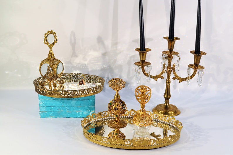 Puede incluir: Un conjunto de tres bandejas decorativas doradas con detalles ornamentados. Las bandejas presentan superficies espejadas y son perfectas para exhibir objetos peque&ntilde;os o joyas. Las bandejas est&aacute;n dispuestas sobre una superficie blanca con un candelero dorado en el fondo.