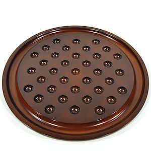 May include: A round wooden solitaire game board with a dark brown finish. The board features a series of evenly spaced holes, each designed to hold a marble. The board has a raised edge and a smooth, polished surface.