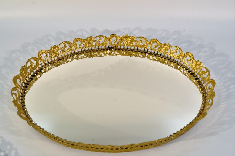 Puede incluir: Una bandeja ovalada de color dorado con un borde decorativo, adornado y de filigrana. La bandeja tiene una superficie de espejo.